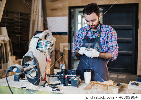 carpenter texting someone on his smart phone near circular saw in a dusty workshop carpenter texting someone on his smart phone near circular saw in a dusty workshop 40899964