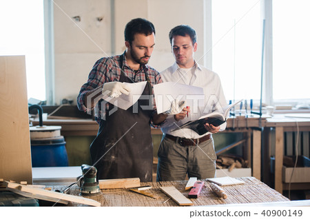 Happy male carpenter showing something to coworker at his notebook papers in workshop 40900149