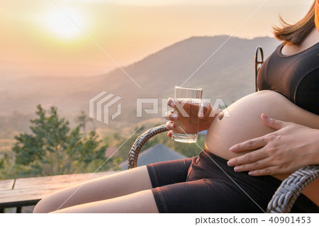 Pregnant woman holding drink water glass on belly on a chair at Pregnant woman holding drink water glass on belly on a chair at 40901453