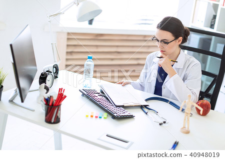 A beautiful young girl in a white robe is sitting at a computer desk with documents and a pen in her 40948019