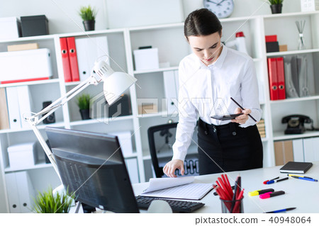 A young girl in the office is standing near the table, holding a pencil and a calculator in her hand 40948064