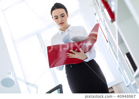 Young girl in the office near the rack and scrolls through the folder with the documents. 40948067