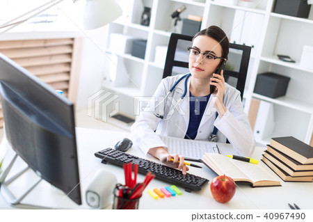 A beautiful young girl in a white robe is sitting at the table, talking on the phone and holding her 40967490