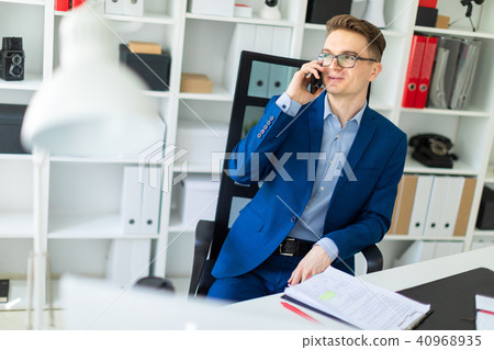 A young man is sitting at a table in the office and talking on the phone. A young man is sitting at a table in the office and talking on the phone. 40968935