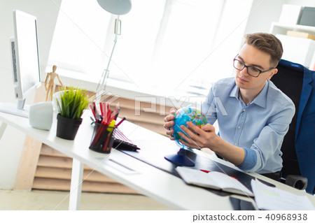 A young man sits in the office at a computer desk and holds a globe in his hands. A young man sits in the office at a computer desk and holds a globe in his hands. 40968938