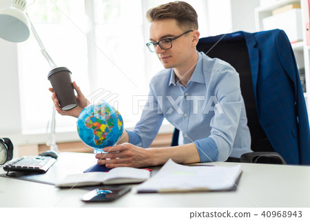 A young man sits in the office at a computer desk and holds a glass of coffee in his hand. A young A young man sits in the office at a computer desk and holds a glass of coffee in his hand. A young 40968943