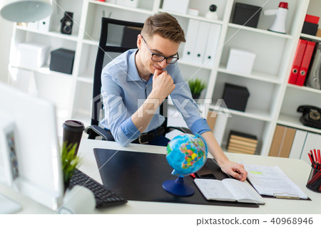 A young man sits in the office at a computer desk and in front of him stands a globe. A young man sits in the office at a computer desk and in front of him stands a globe. 40968946