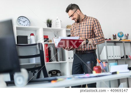 A man in the office stands near the shelf and scrolls through the folder with the documents. 40969147