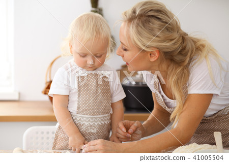 Little girl and her blonde mom in beige aprons  playing and laughing while kneading the dough in th 41005504