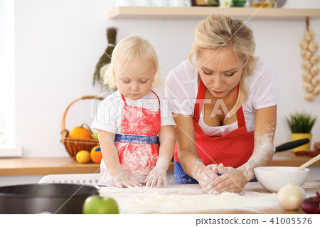 Little girl and her blonde mom in red aprons playing and laughing while kneading the dough in th Little girl and her blonde mom in red aprons playing and laughing while kneading the dough in th 41005576