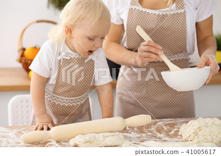 Little girl and her blonde mom in beige aprons playing and laughing while kneading the dough in th Little girl and her blonde mom in beige aprons playing and laughing while kneading the dough in th 41005617