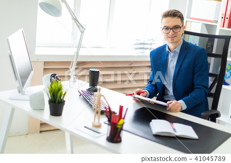 A young man sits at a table in the office and holds documents and a pen in his hands. 41045789