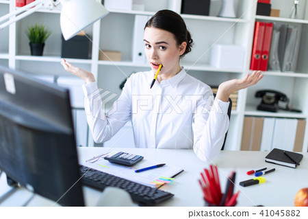 A young girl in the office sits at a table, holding a pen in her mouth and spreading her hands to 41045882
