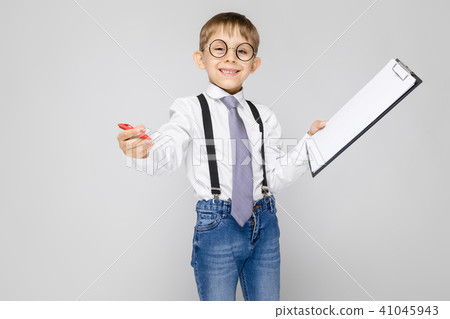 A charming boy in a white shirt, suspenders, a tie and light jeans stands on a gray background. The 41045943