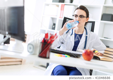 A beautiful young girl in a white robe sits at a computer desk, drinks water and looks at the 41046686