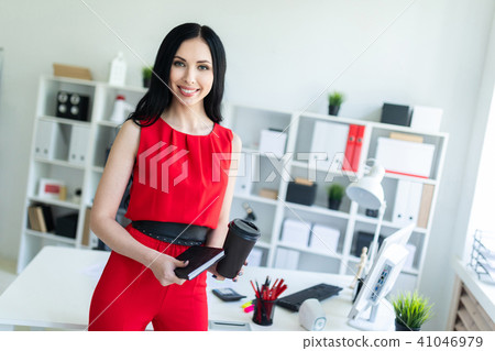 Beautiful young girl in a red suit is standing in the office and is holding a notebook and a glass Beautiful young girl in a red suit is standing in the office and is holding a notebook and a glass 41046979