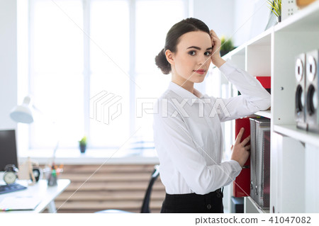A young girl in the office rests her hand on the shelf and pulls out a red folder. 41047082