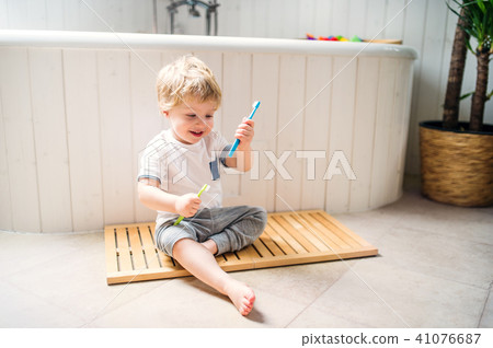 A toddler boy with a toothbrush sitting on the floor in the bathroom at home. 41076687