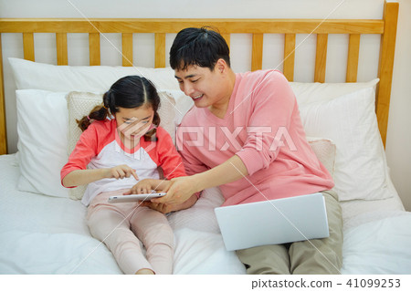 一個場景，其中一個父親和女兒在床上愉快地休息，同時玩一台筆記本電腦和一個墊 41099253