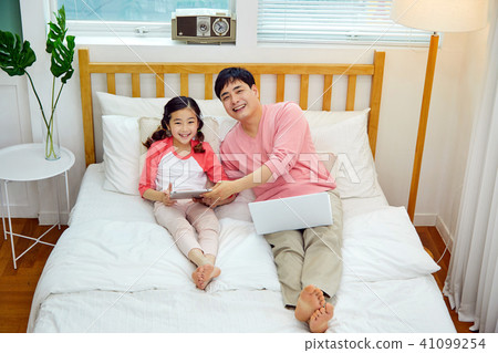 一個場景，其中一個父親和女兒在床上愉快地休息，同時玩一台筆記本電腦和一個墊 41099254