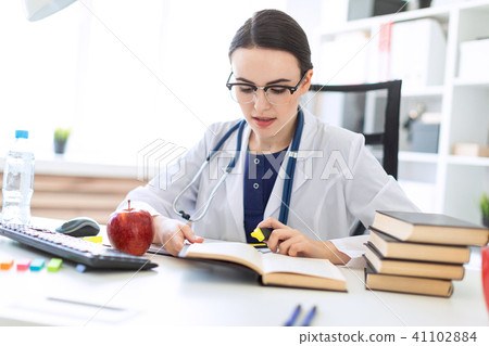 A beautiful young girl in a white robe is sitting at the computer desk with a marker and a book. A 41102884