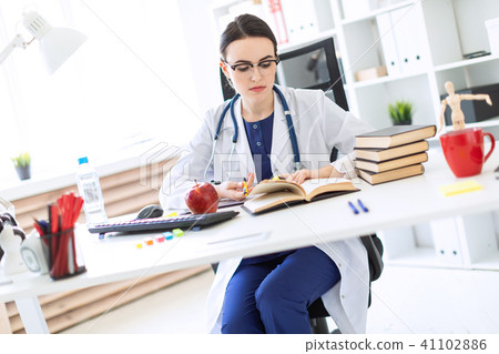 A beautiful young girl in a white robe sits at a computer desk, holds a marker and a pen in her hand 41102886