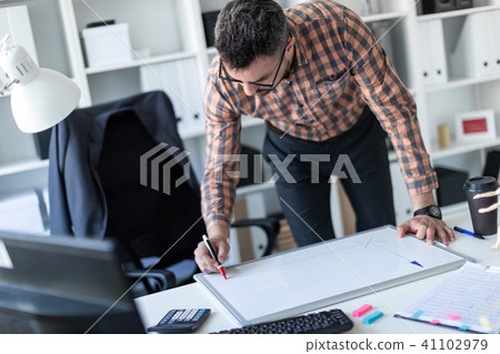 A man in the office is standing near the table and draws a marker on the magnetic board. 41102979