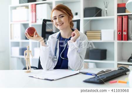 A beautiful young girl in a white robe is sitting at a table in the office and is holding an apple A beautiful young girl in a white robe is sitting at a table in the office and is holding an apple 41103068