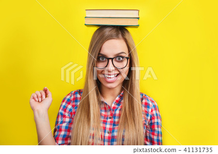 young woman in glasses holding books on her head. Smiling young stylish student is standing with young woman in glasses holding books on her head. Smiling young stylish student is standing with 41131735