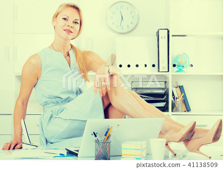 young cheerful adult girl in blue dress sitting on table in home office 41138509