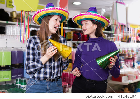 Attractive girls trying on party hats in festive accessories store Attractive girls trying on party hats in festive accessories store 41146194