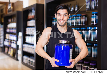 Young bodybuilder showing power and holding pot of sport nutrition products in shop Young bodybuilder showing power and holding pot of sport nutrition products in shop 41147685
