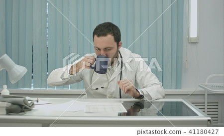 tired from work, bearded doctor drink from cup and continue woking with documents and x-rays tired from work, bearded doctor drink from cup and continue woking with documents and x-rays 41180427