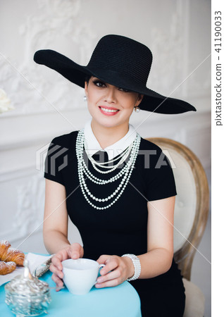 Attractive woman wearing black dress, hat and pearls, sitting on chair Attractive woman wearing black dress, hat and pearls, sitting on chair 41190033