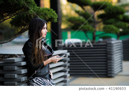 Beautiful young dark-haired woman is texting on her smartphone while sitting the bench in city park Beautiful young dark-haired woman is texting on her smartphone while sitting the bench in city park 41190933