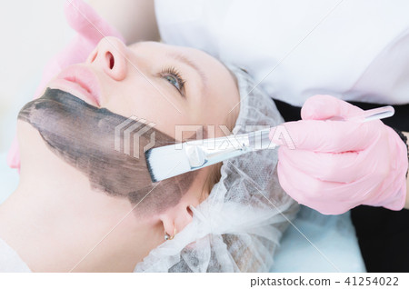 The cosmetologist in pink gloves with a brush applies a carbon mask for peeling on the face of a The cosmetologist in pink gloves with a brush applies a carbon mask for peeling on the face of a 41254022