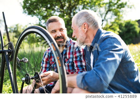 An adult hipster son and senior father repairing bicycle outside on a sunny day. 41255889