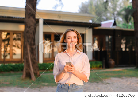 Young blonde woman chatting by smarptone near resort house, wearing blouse and shorts. 41263760