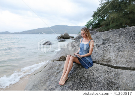 Young woman working with laptop on sea shore and sitting on stone. Young woman working with laptop on sea shore and sitting on stone. 41263817