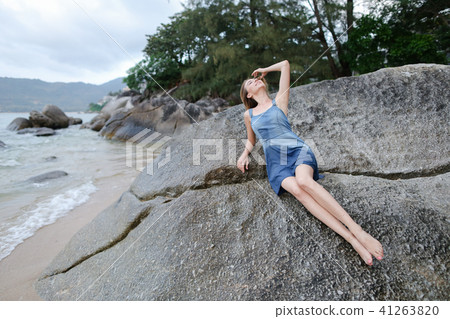 Young caucasian woman lying on stones on sea shore in morning, wearing jeans sundress. 41263820