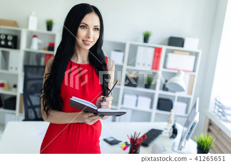 Beautiful young girl in a red suit is standing in the office and holding a notebook and a pencil. Beautiful young girl in a red suit is standing in the office and holding a notebook and a pencil. 41265651