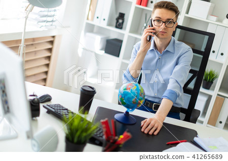 A young man is sitting in the office at a computer desk and talking on the phone. Before the young 41265689