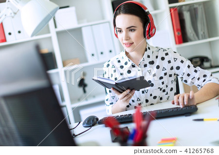 Beautiful young girl sits in headphones at desk in office, holds a notebook in her hand and prints 41265768