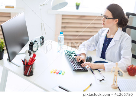 A beautiful young girl in a white robe is sitting at the table and typing on the keyboard. 41265780