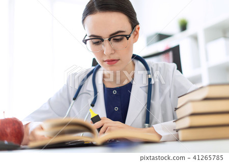 A beautiful young girl in a white robe is sitting at the computer desk with a marker and a book. A 41265785