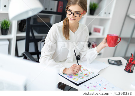 A young girl is sitting at a computer desk in the office, holding a pencil and a red cup. Before the A young girl is sitting at a computer desk in the office, holding a pencil and a red cup. Before the 41298388