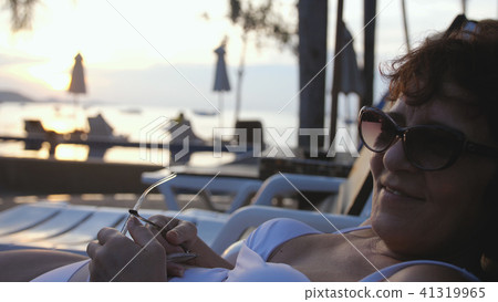 Woman in aged wearing sunglasses and bikini relaxing in resort by the beach in sunbed. 41319965
