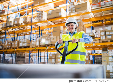 A senior woman warehouse worker pulling a pallet truck with boxes. A senior woman warehouse worker pulling a pallet truck with boxes. 41321760