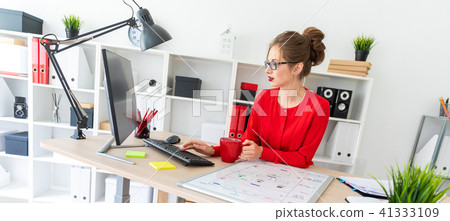 A young girl is sitting at the desk in the office, holding a red cup in her hand and typing text on 41333109