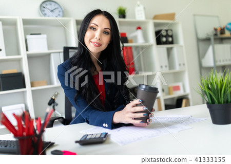 A beautiful young girl is holding a glass with coffee, sitting on a chair in the office at the table 41333155